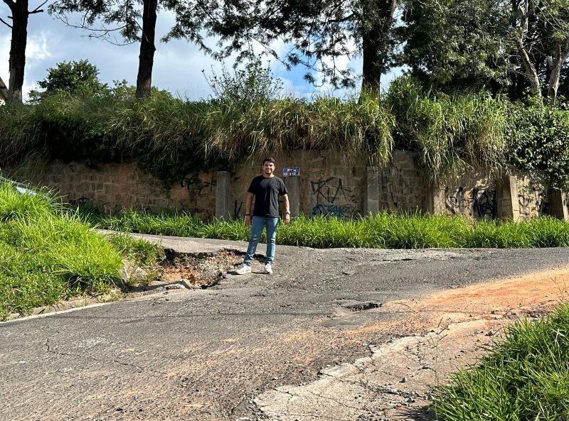 Vereador Rafael Tanzi em frente a trecho danificado de via pública. Estrutura de muro e vegetação completam a imagem.