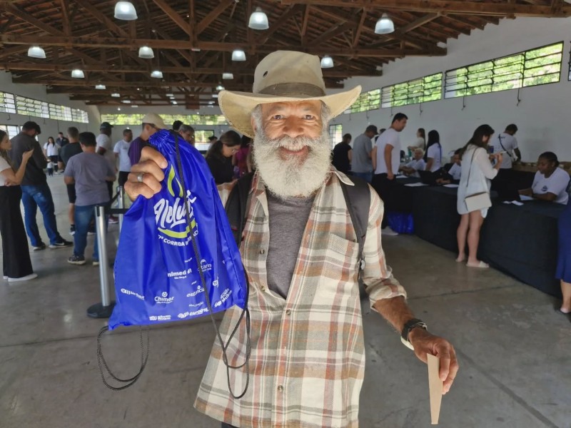 Um participante sorri ao exibir o kit da Corrida de Aleluia. Ao fundo, pessoas aguardam a distribuição dos kits, em evento.