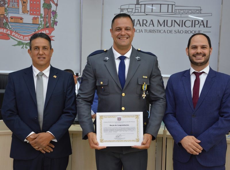 Leandro da Silva Santos, em uniforme militar, recebe homenagem de vereadores. Ele segura certificado ladeado por dois homens em trajes sociais, sorrindo. Cena ocorre em Câmara Municipal.