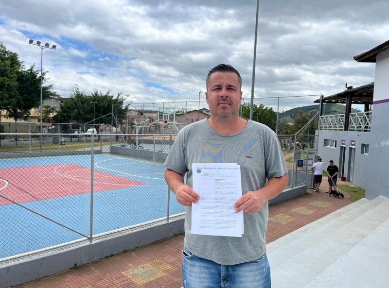Vereador Thiago Nunes segura documentos, em frente a quadra esportiva. Ele solicita transporte e uniformes para alunos do programa Jogando Junto.