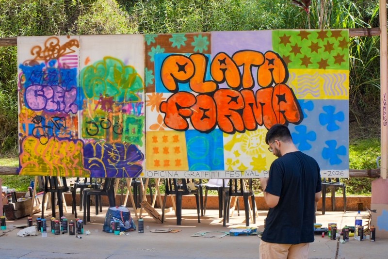 Artista finaliza grafite com a palavra "PLATAFORMA" em painel. Latas de spray e materiais de pintura estão dispostos no chão, indicando oficina em festival de cultura urbana.