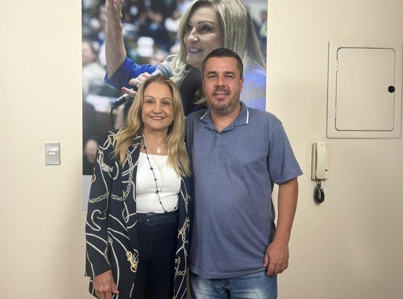 O vereador Thiago Nunes posa com outra pessoa em frente a um banner. A imagem é de dentro de um escritório, com detalhes na parede.