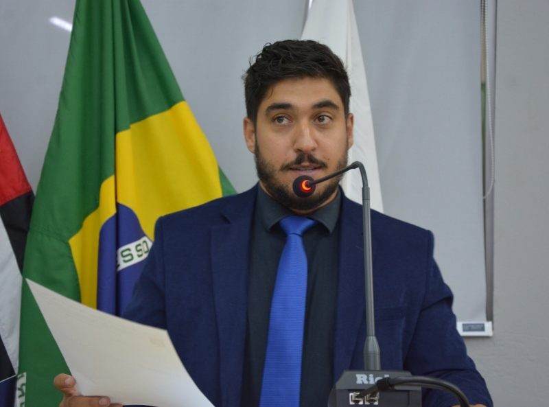 Vereador Diego Costa discursa em evento, segurando documentos. Ao fundo, bandeiras do Brasil e do estado.