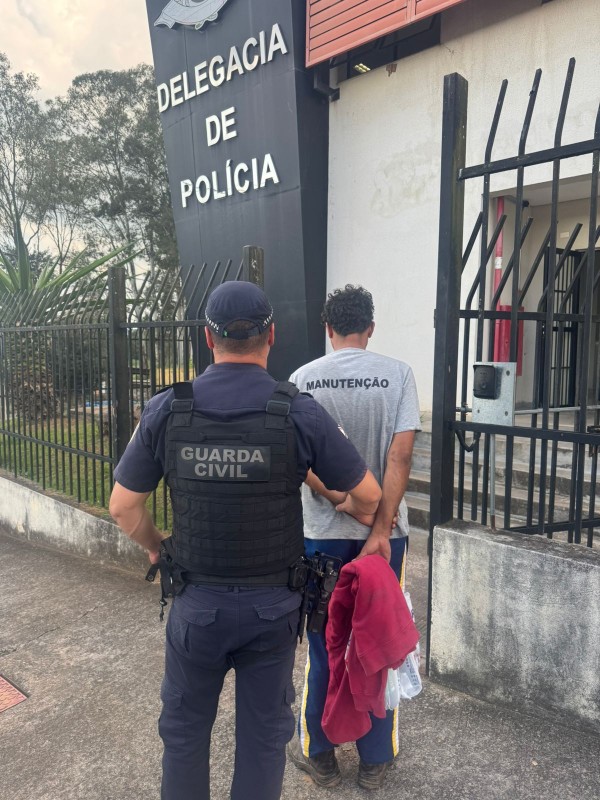 Um agente da Guarda Civil conduz um homem algemado em frente à delegacia. O preso usa camiseta "Manutenção".