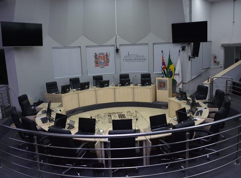 Plenário da Câmara Municipal, com mesas e cadeiras dispostas em semicírculo. No fundo, painéis e bandeiras. Imagem ilustra o anúncio do encerramento da sessão.