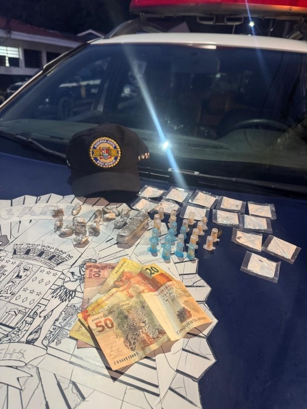 Em cima de um capô, estão drogas, seringas, embalagens, dinheiro e um boné da Guarda Municipal. A apreensão ocorreu em São João Novo.