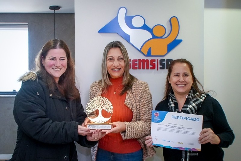 Equipe de São Roque celebra prêmio por projeto inovador em saúde mental. Na foto, mulheres exibem troféu e certificado em ambiente institucional. Logotipo da EMS SP ao fundo.