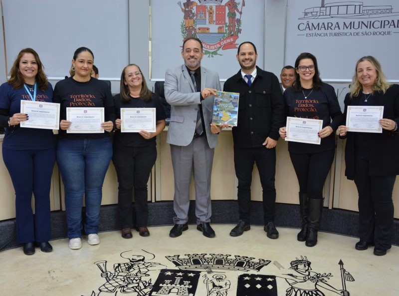 Em São Roque, Wanderlei da Qualiser posa com equipe do SUAS, após homenagem. Servidores seguram diplomas e livro, em frente à Câmara Municipal.