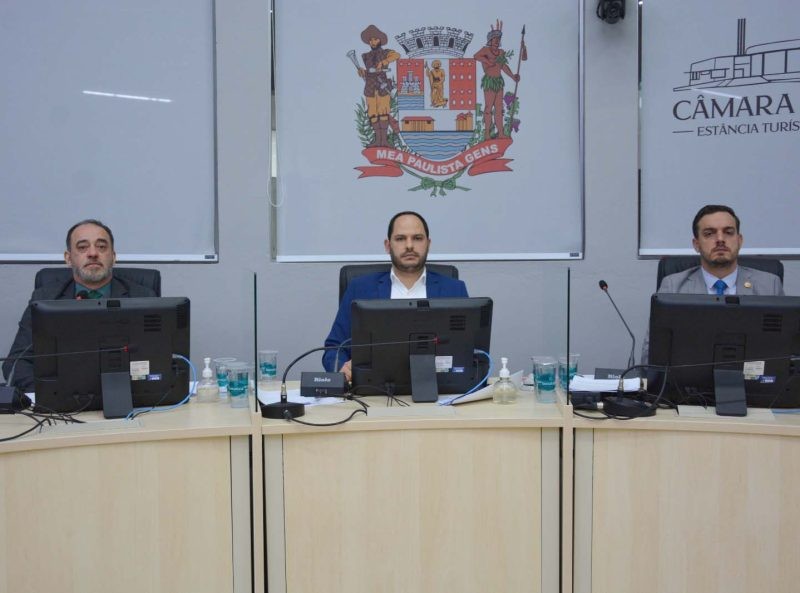 Membros de comissão parlamentar sentados em mesa de reunião, com computadores e microfones. Ao fundo, brasão e identificação da Câmara.