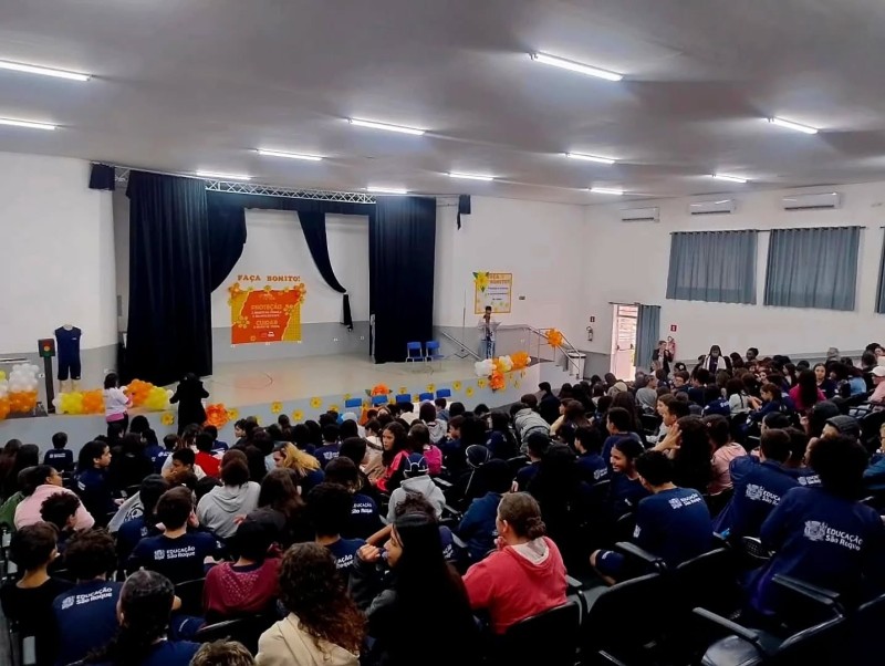 Auditório lotado durante evento de conscientização sobre exploração sexual infantil. No palco, banner e decoração para a data.