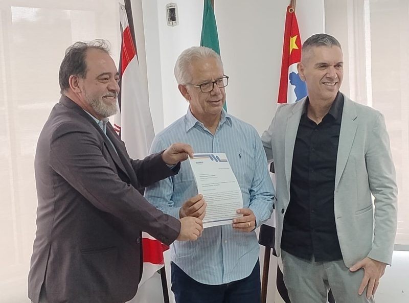 Wanderlei da Qualiser e outros dois homens posam segurando um documento. Ao fundo, bandeiras e um ambiente institucional.