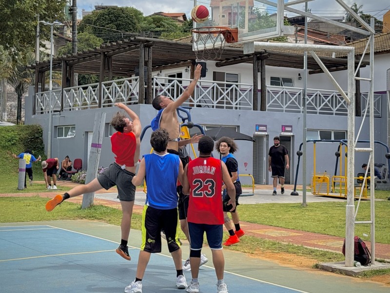 Jogadores disputam lance em partida de basquete. A bola está próxima à cesta, enquanto atletas se movimentam em quadra. A cena se passa em área externa.